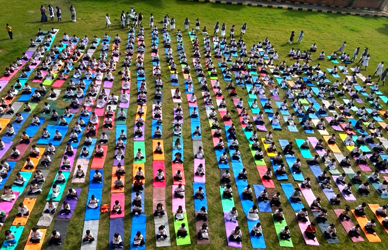 Hundreds of students seated on coloured mats in rows across a lawn at a mass yoga event