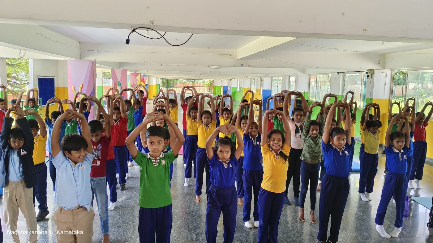 A hall full of school children in uniform with arms raised in a yoga pose